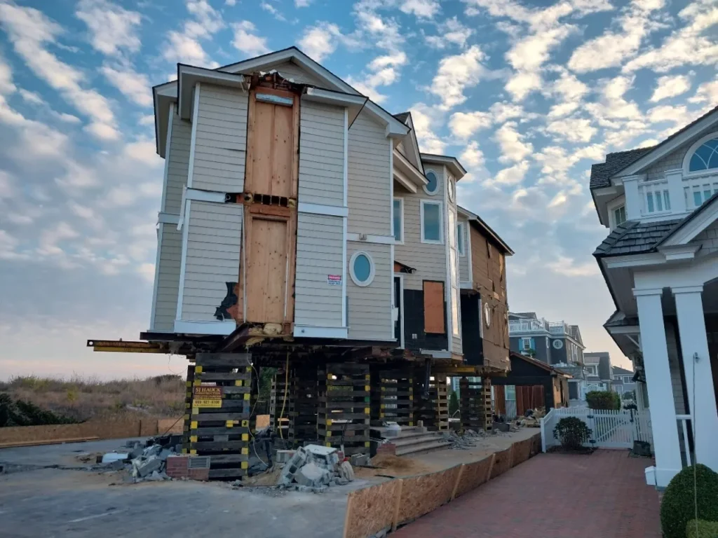 A house elevated on stilts with boarded windows and an unfinished exterior, surrounded by construction debris and a cloudy sky.