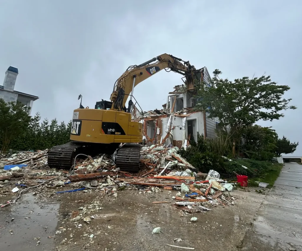 A yellow excavator demolishes a partially destroyed house surrounded by debris on a rainy day.