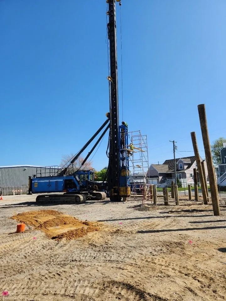 A construction site featuring a tall drilling rig, workers on a scaffold, and wooden posts being installed in the ground.