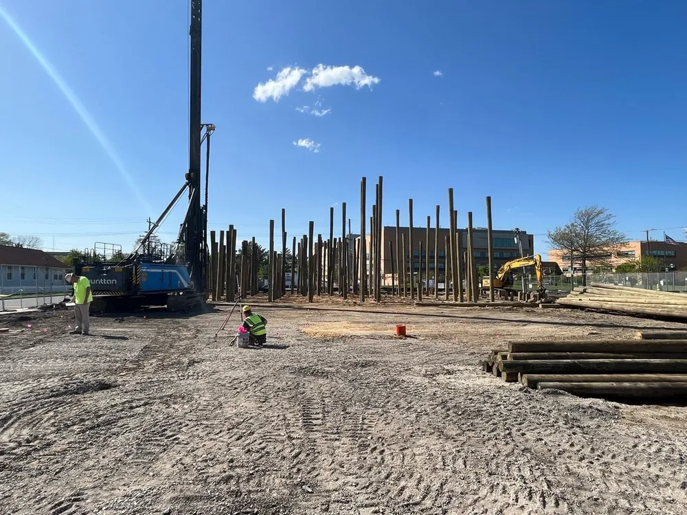 Construction site with vertical wooden poles in the background, a crane on the left, and workers in safety vests on the ground.