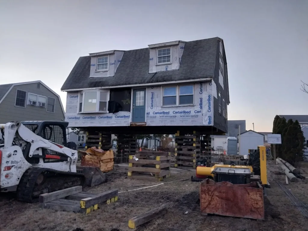 A house raised on stilts at a construction site, with a construction vehicle nearby.