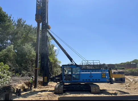 A large blue construction machine with a drilling rig is positioned on a construction site, surrounded by trees and earth. Two workers are visible in safety gear.
