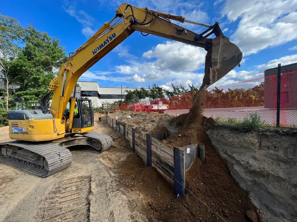 A yellow excavator digging and pouring dirt at a construction site with a red fence in the background under a blue sky.
