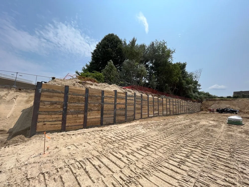 A wooden retaining wall in a construction site, with sandy ground and some trees in the background under a clear blue sky.