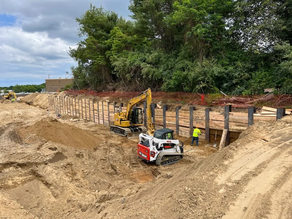 Heavy machinery working on a construction site with excavated earth and wooden supports. Trees and a cloudy sky are visible in the background.