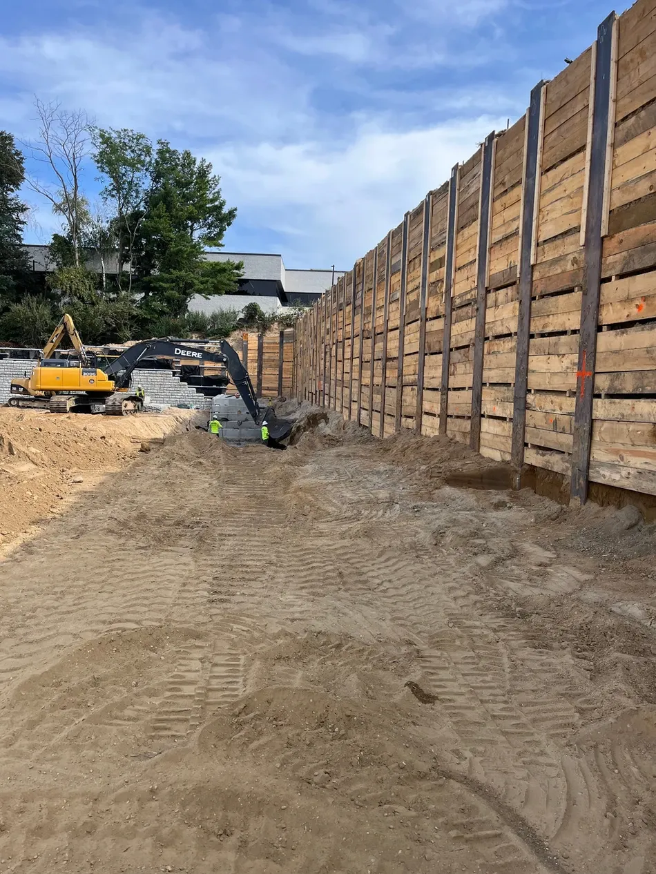 A construction site with a trench surrounded by wooden retaining walls, and construction machinery in the background.
