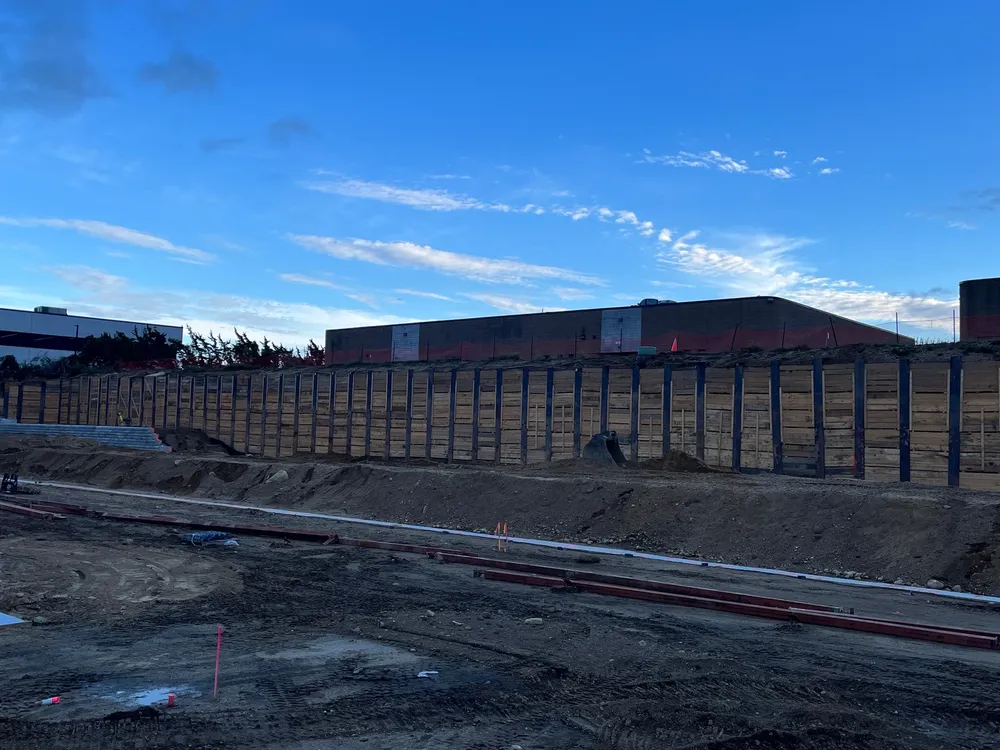 A construction site with a long, vertical wooden or metal wall structure, under a blue sky with some clouds.