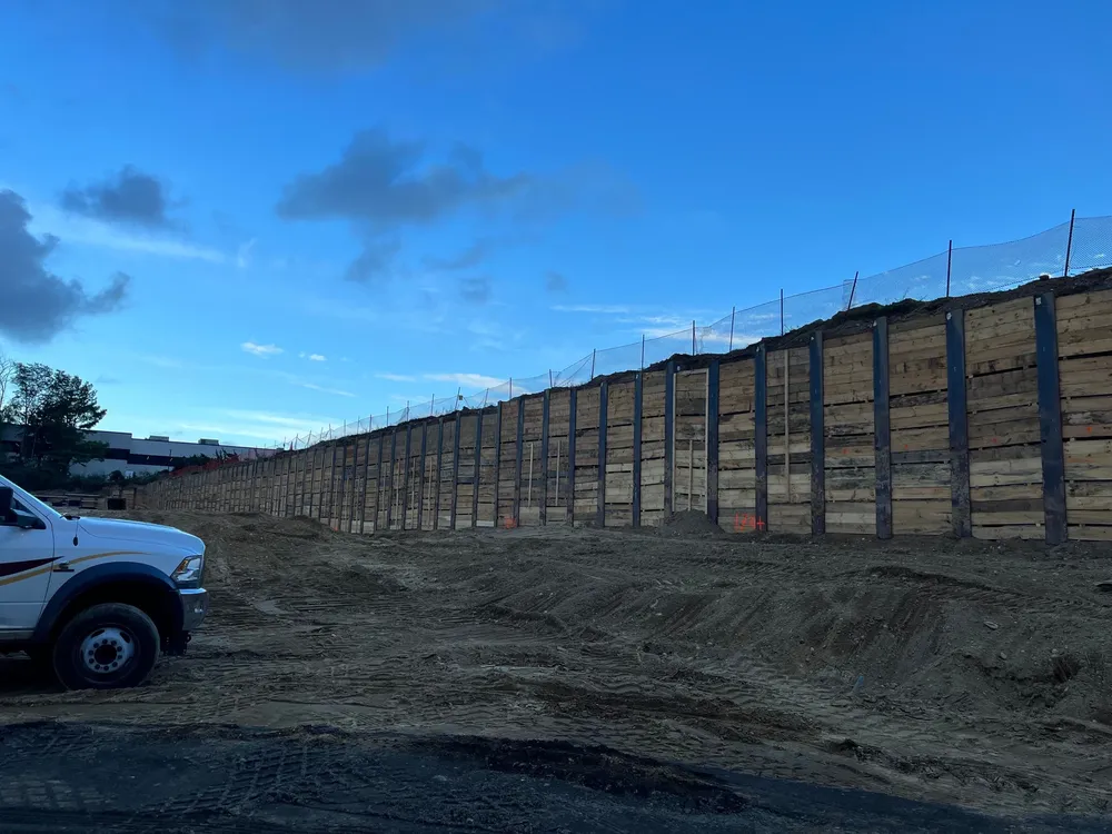 Construction site with a tall wooden retaining wall under a blue sky, with a truck parked nearby.
