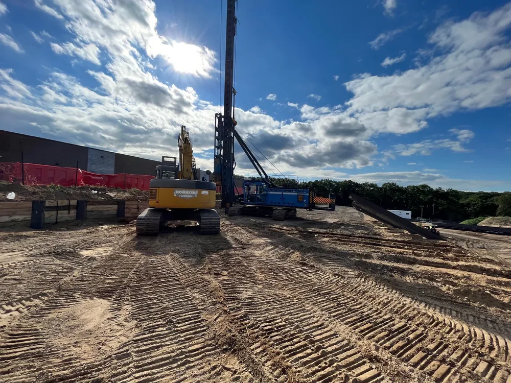 Construction site with heavy machinery, including an excavator and a drilling rig, against a cloudy sky.