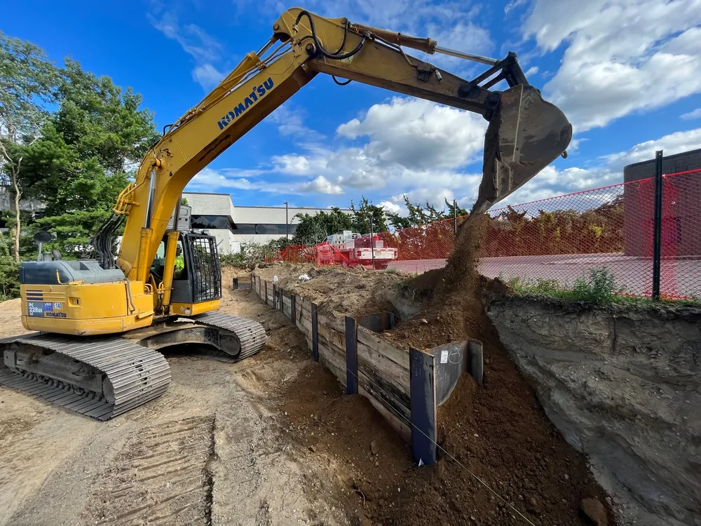 An excavator digging and moving soil at a construction site, with a blue sky and clouds in the background.