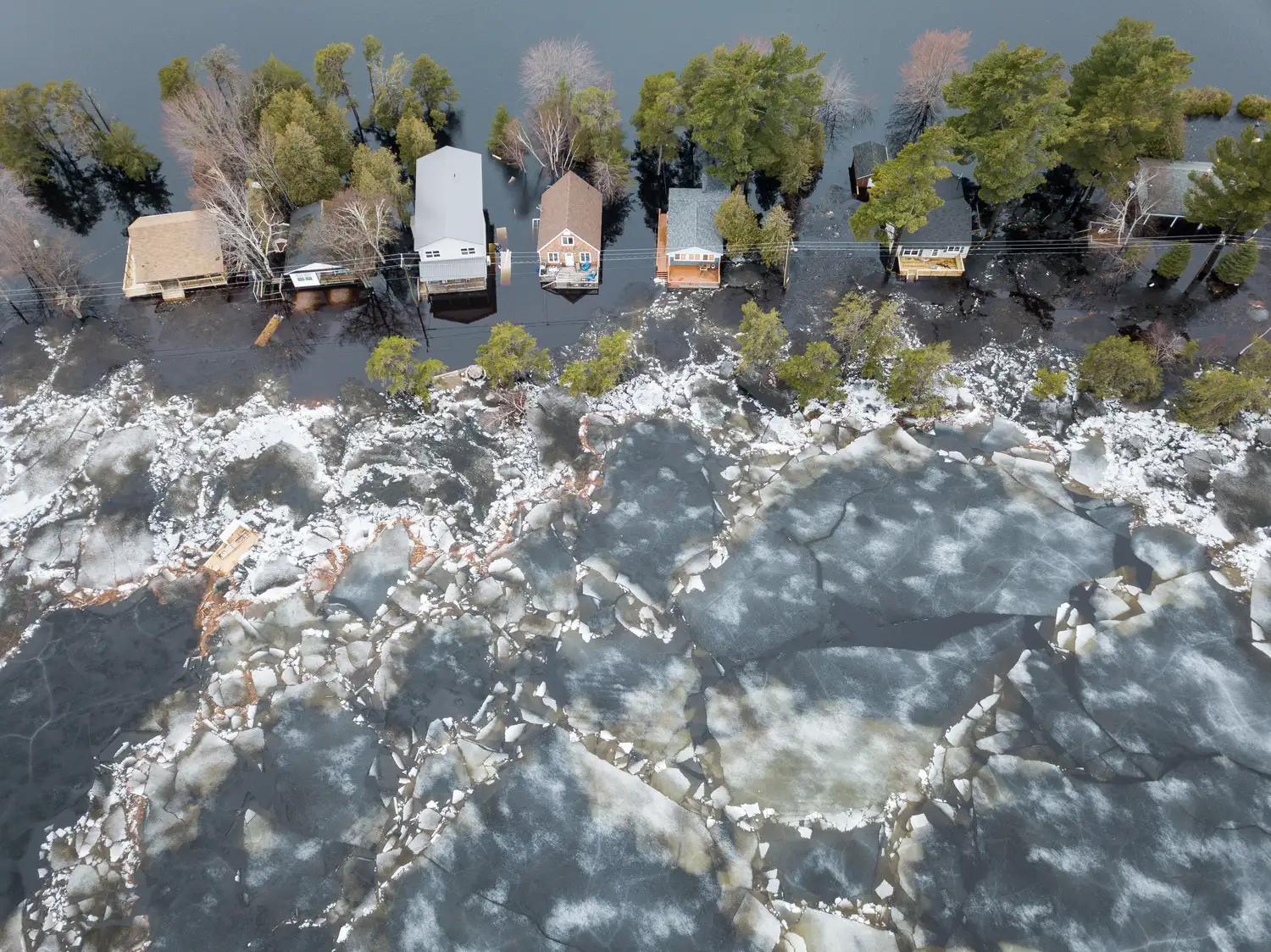 An aerial view of houses near a flooded area, with water surrounding the buildings and ice formations in the foreground.