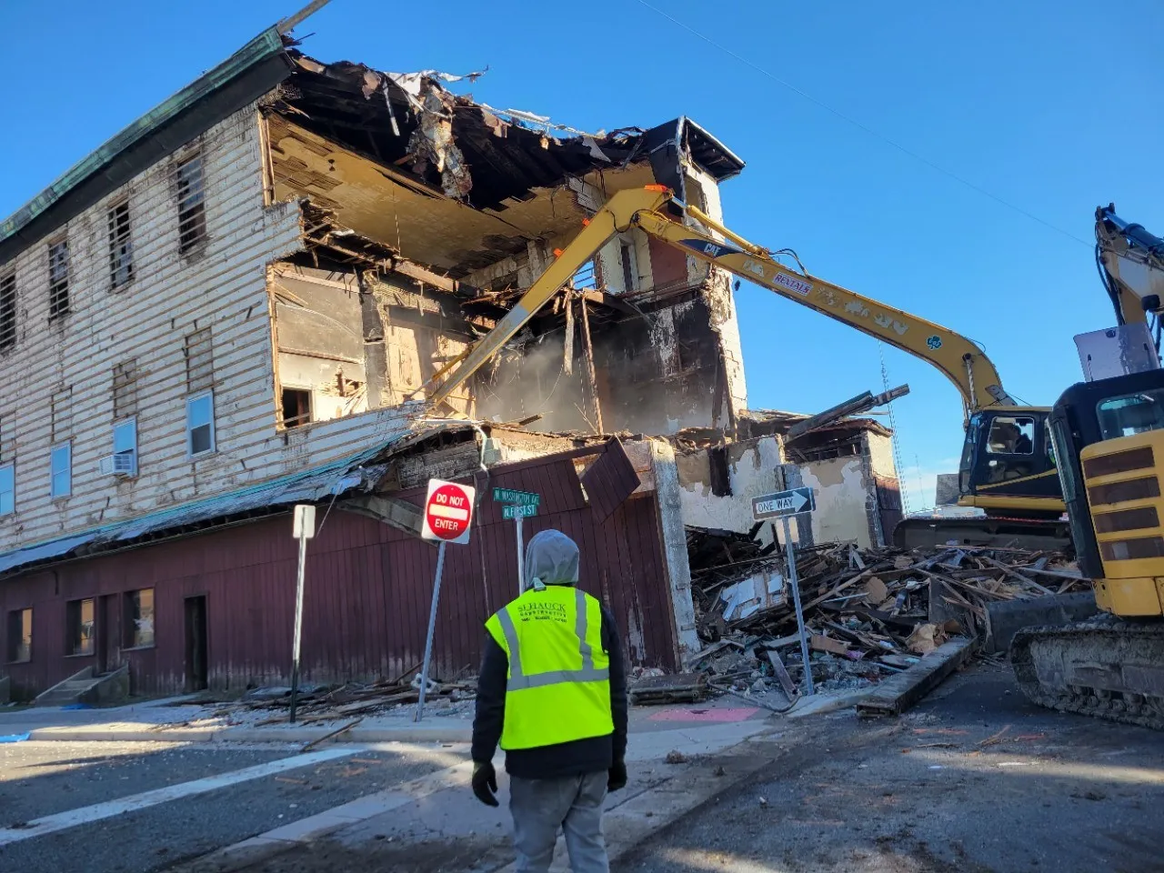 A construction worker in a yellow safety vest observes a building being demolished by heavy machinery, with debris scattered around and a clear blue sky above.