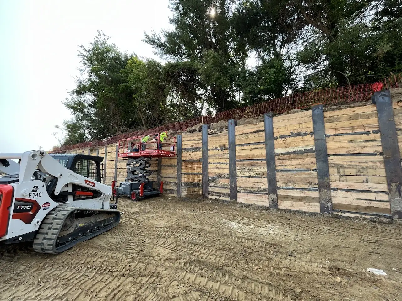 Construction site with heavy machinery, including a skid steer loader and a scissor lift, near a wooden wall structure.