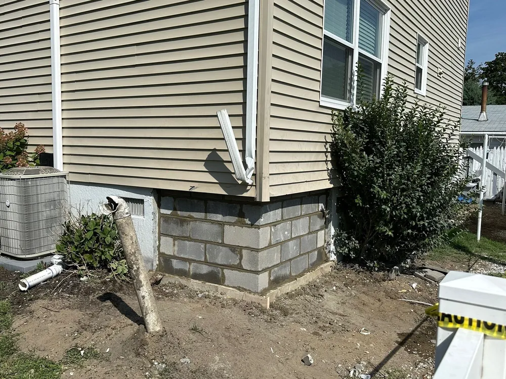 A corner of a house showing a foundation made of concrete blocks, with a downspout and a bush nearby; the ground is bare with some construction debris and a partially visible air conditioning unit.
