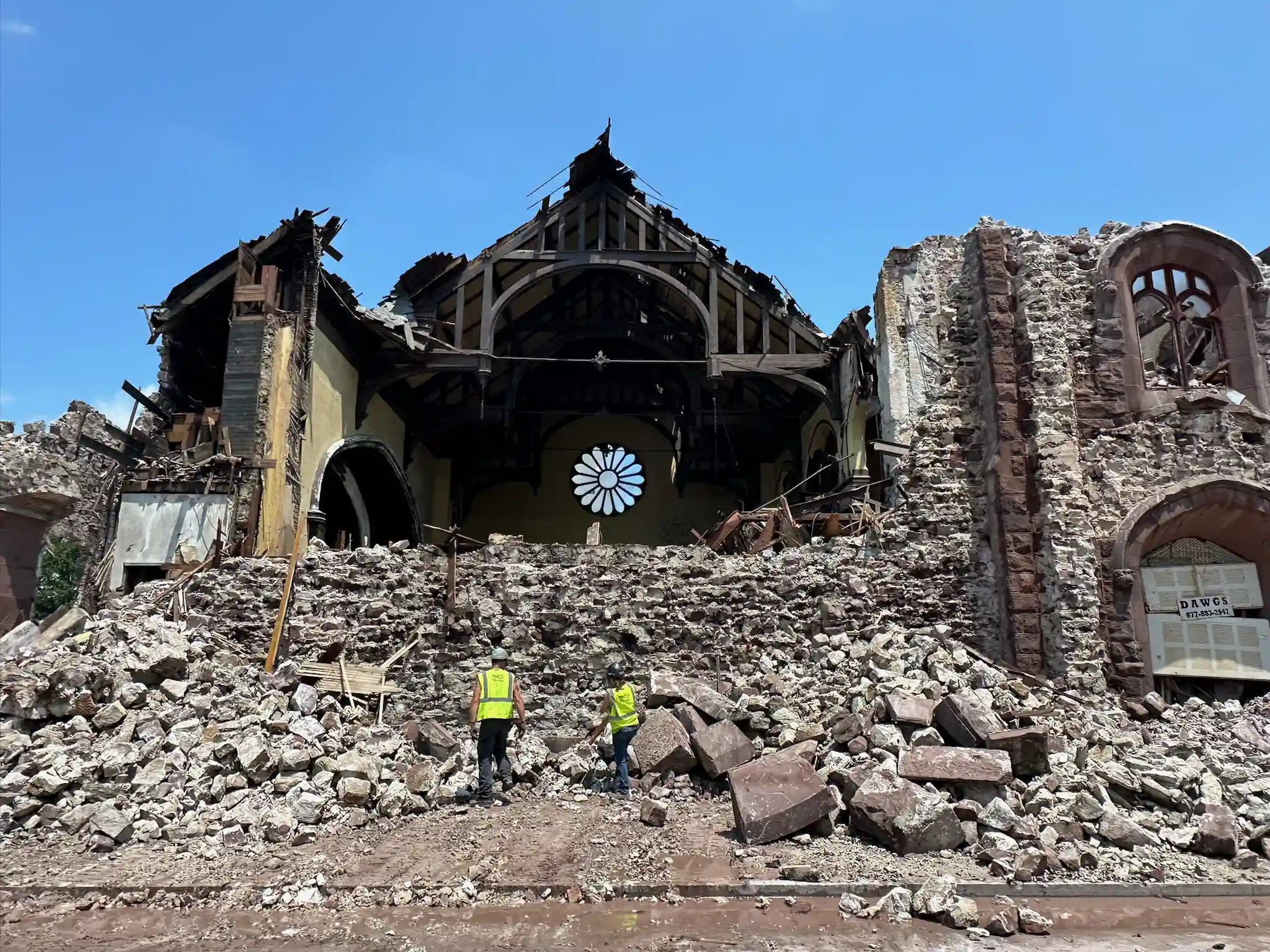 A partially collapsed building with a large round window, surrounded by rubble, and two people in safety vests standing in front.