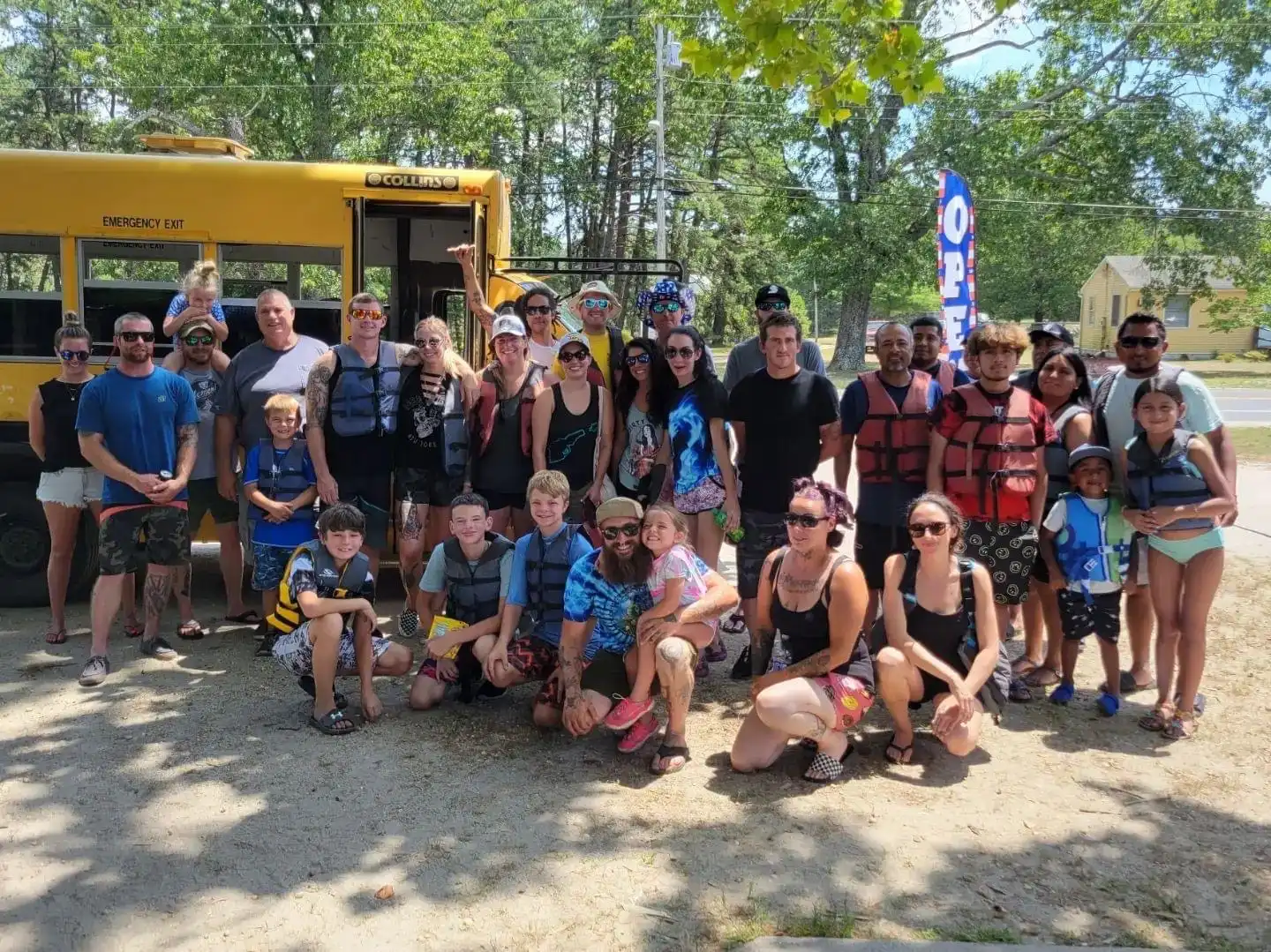 A large group of people, including children and adults, posing together in a sunny outdoor setting near a yellow school bus. Many are wearing life jackets and swim attire.