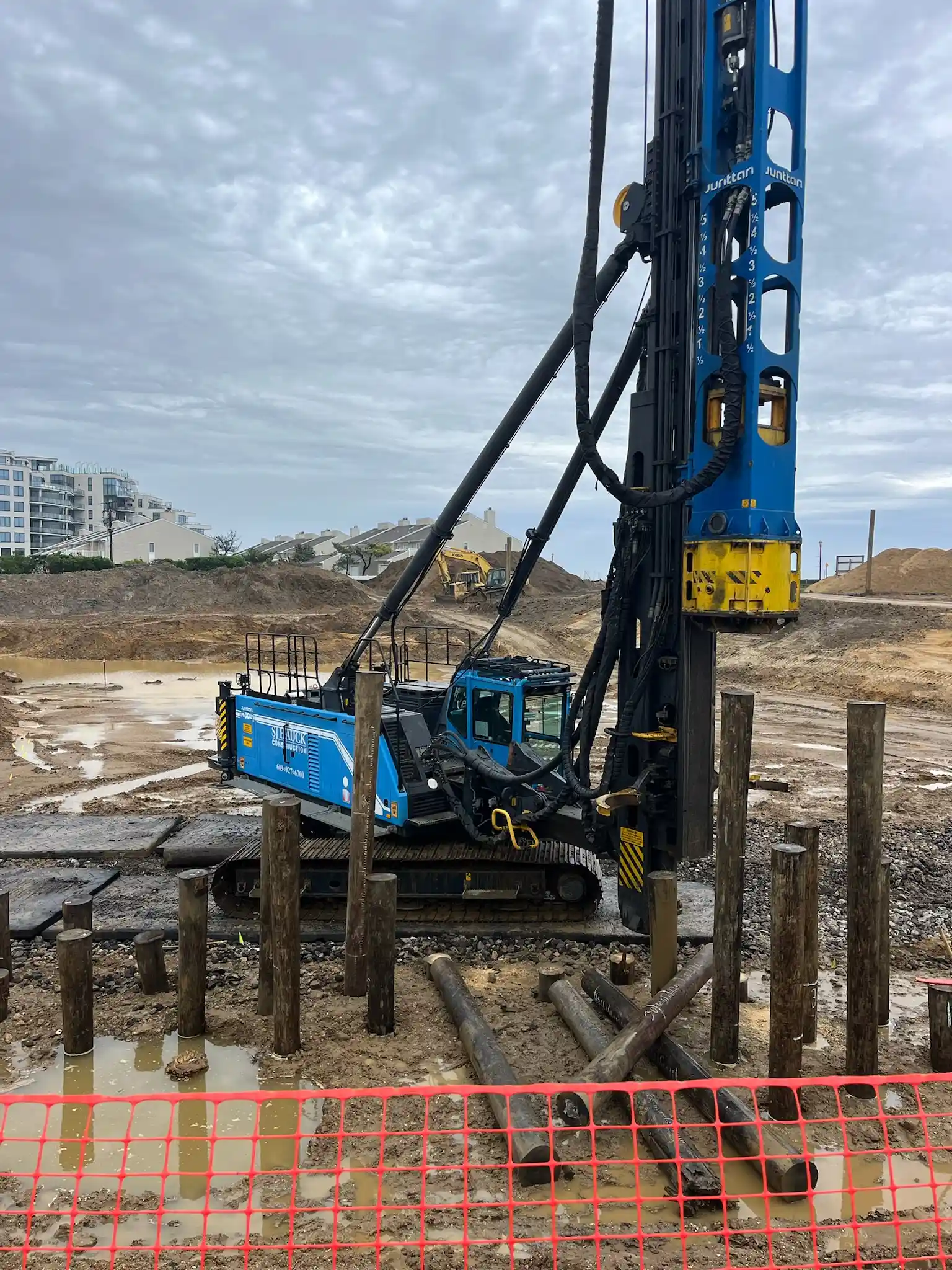 A heavy-duty drilling machine on a construction site, surrounded by wooden piles and muddy ground, with cloudy skies in the background.