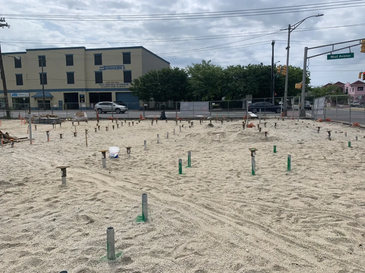 Construction site with sand and concrete pilings in the ground, surrounded by fencing and buildings in the background.