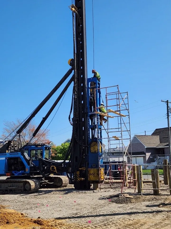 A construction site with a drill rig and workers on scaffolding, set against a clear blue sky.
