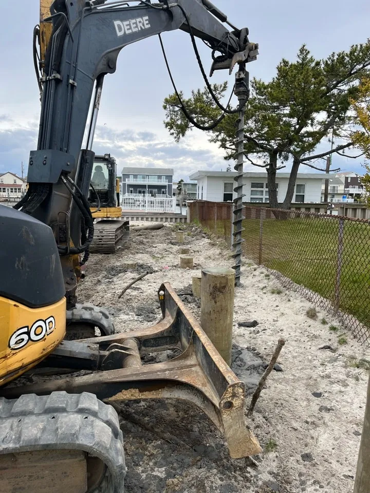 A construction site with a yellow excavator operating a drill, surrounded by a fenced area and trees in the background.