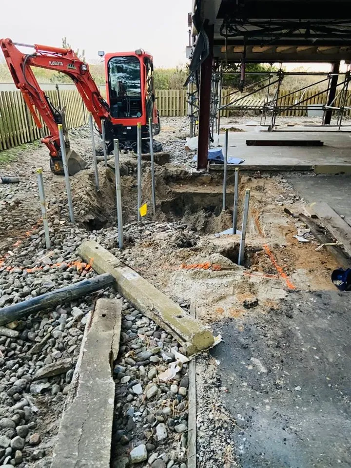 A large yellow tracked excavator with a hydraulic drive attachment installing a steel helical pile foundation into the ground at an active construction site, with an operator visible inside the cab and structural steel building frames in the background.