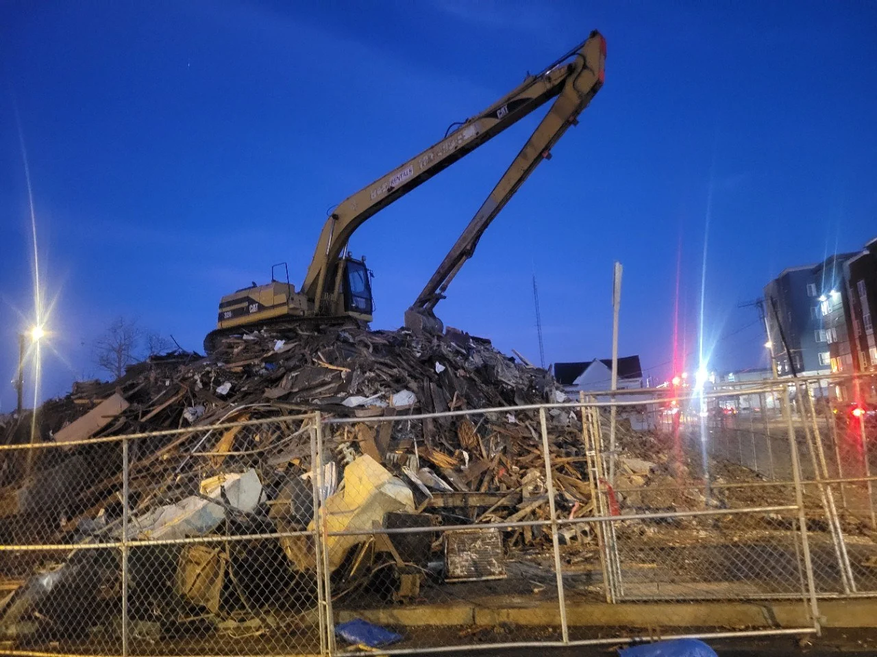 A large construction vehicle is lifting materials from a tall pile of debris at dusk, surrounded by a chain-link fence.