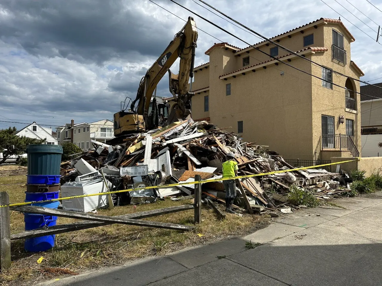 A large excavator is demolishing a pile of debris next to a building, with a person in a neon vest standing nearby.