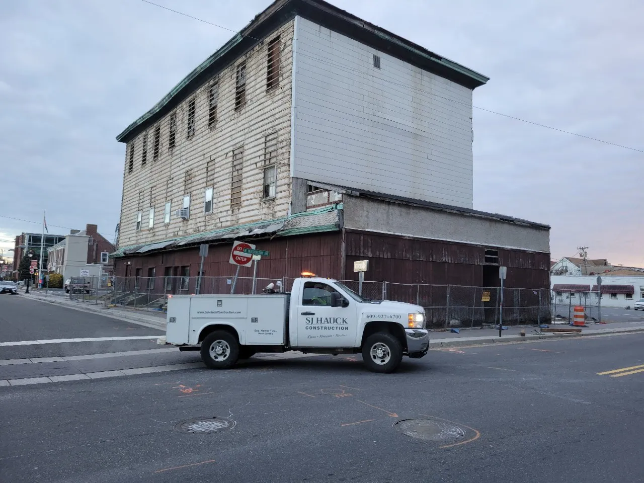 A white pickup truck parked in front of an old, partially boarded-up building under a cloudy sky.