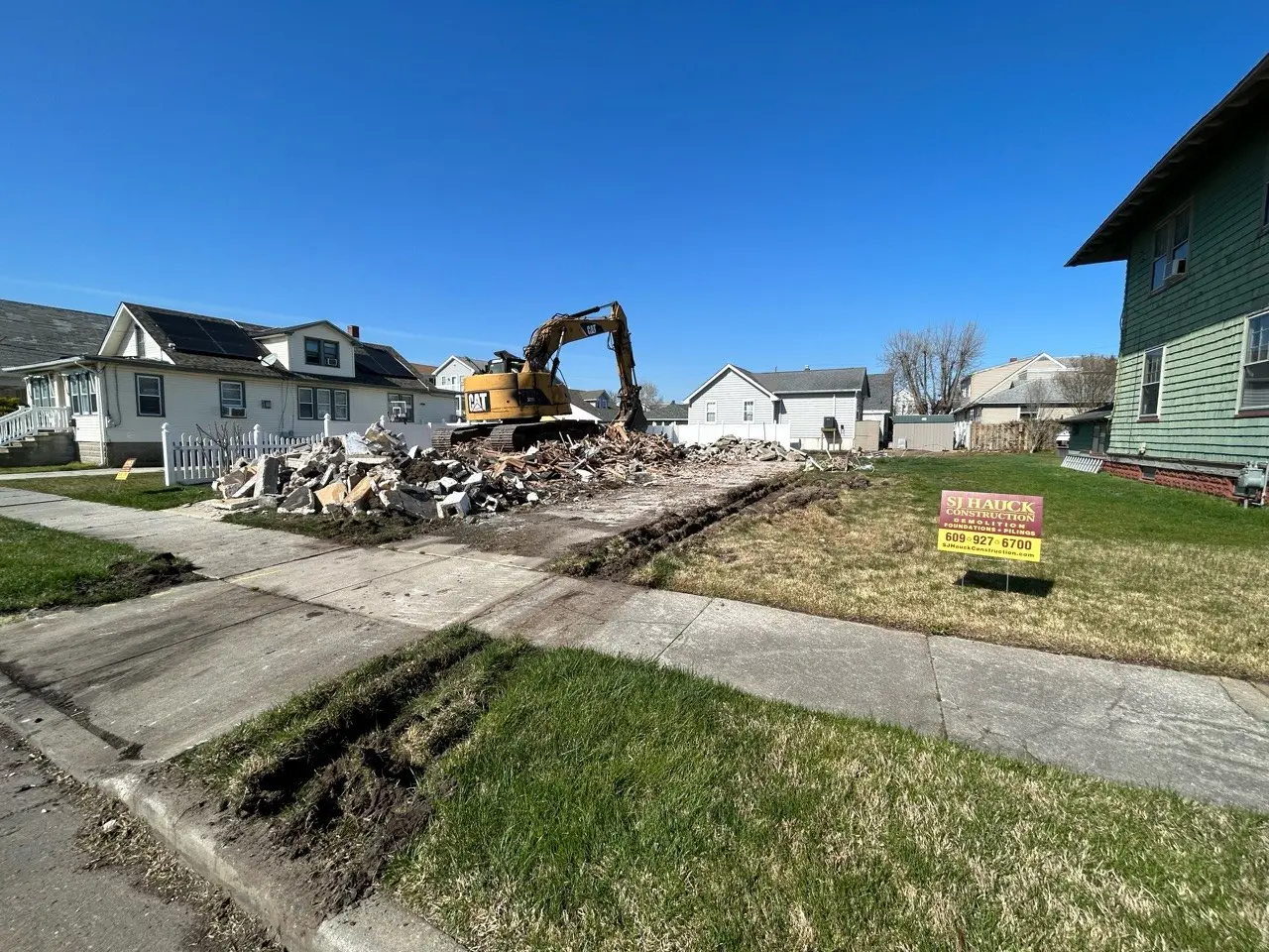 A construction site with machinery working on a demolished area, surrounded by houses and a for sale sign.