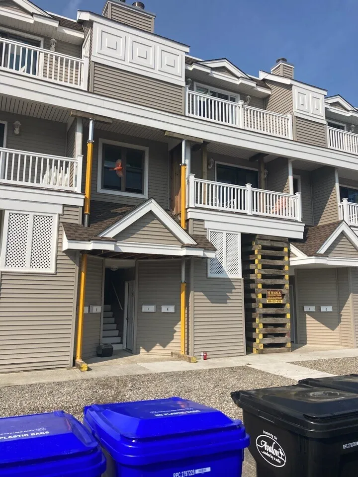 A multi-story apartment building with a wooden staircase leading to the second floor, plastic trash bins in blue and black in the foreground, and a clear blue sky above.