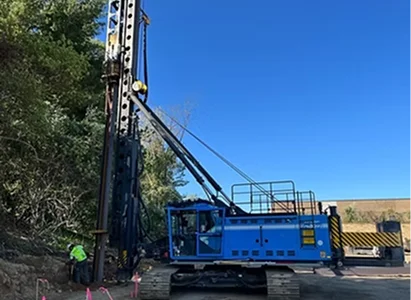 A blue drilling machine is parked on a construction site next to a hill covered in trees.