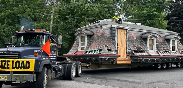 A truck transporting a house, with a construction worker on top, surrounded by trees and power lines.