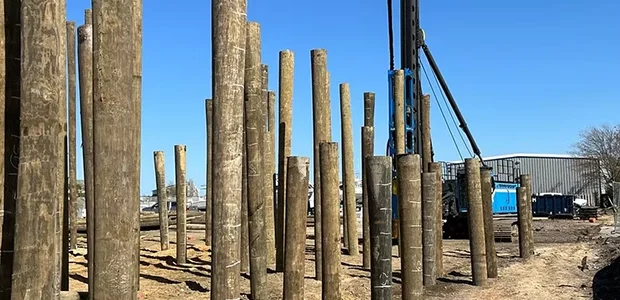 A construction site with several upright wooden poles and machinery in the background against a clear blue sky.