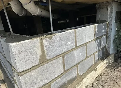 A partially constructed concrete block wall under a structure, showing cement mortar between the blocks and exposed plumbing above.