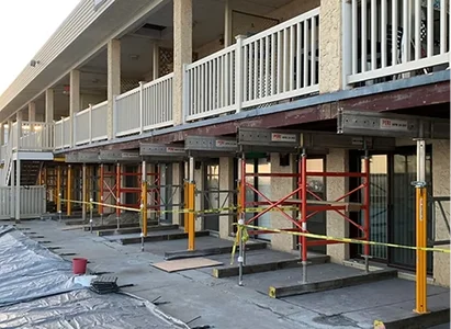 Construction scaffolding set up in front of a building, supporting structures and covered with tarps. Balconies above the scaffolding are visible.