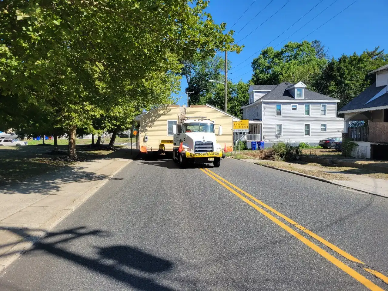 A large truck towing a house down a residential street lined with trees and houses.