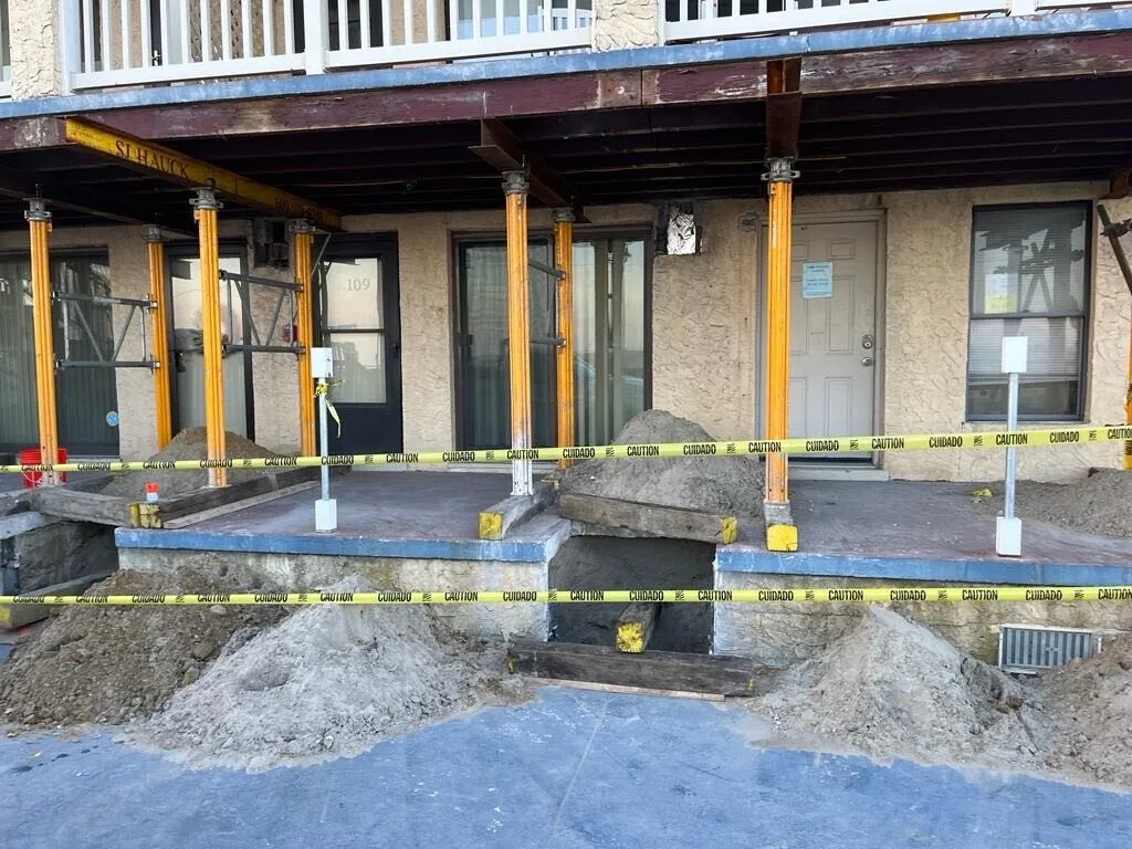 Construction site with yellow caution tape around a building entrance, featuring scaffolding, bare earth, and large rocks.