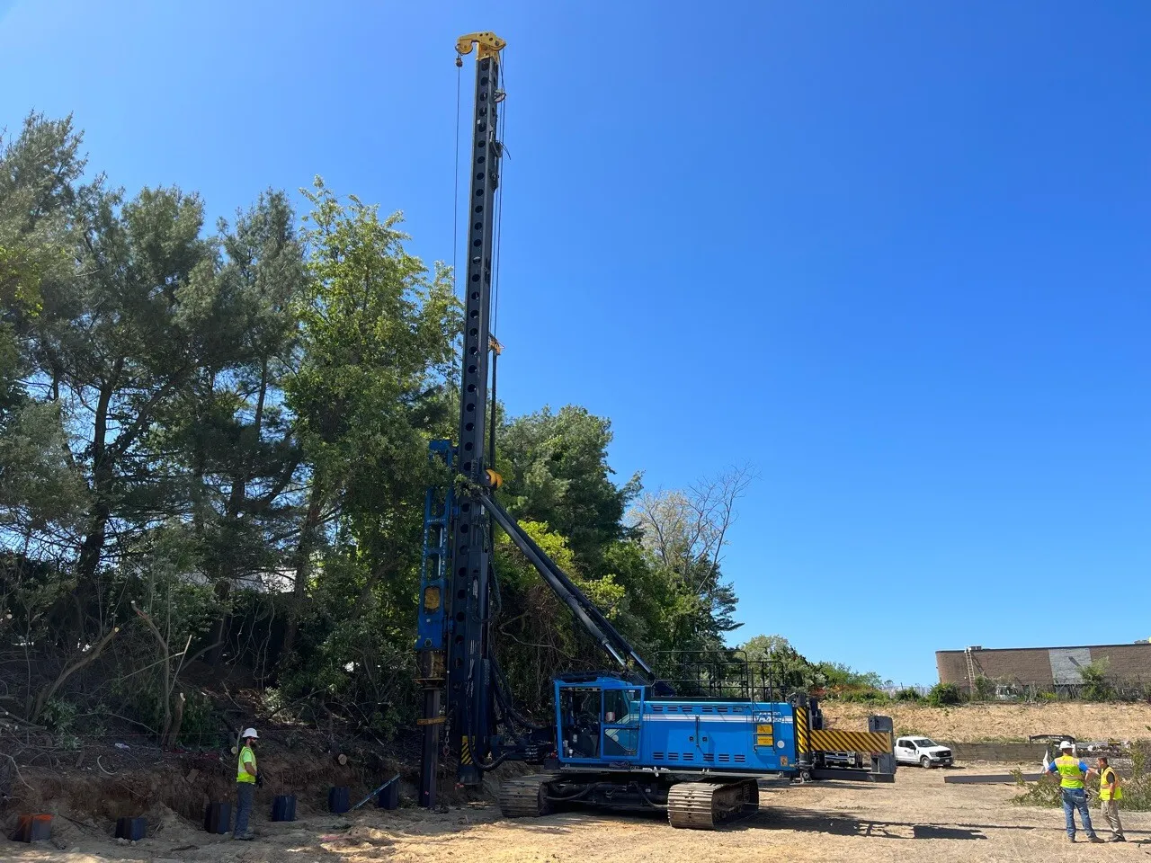 A tall drilling rig next to a wooded area, with workers in safety vests nearby.