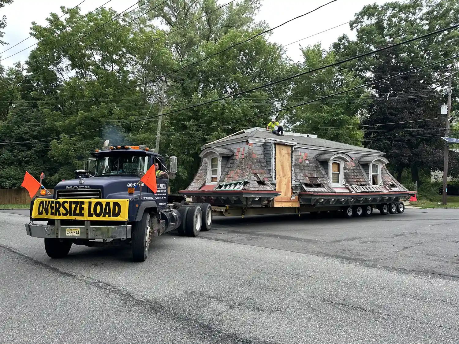 A large truck transporting a house, with an "Oversize Load" sign, driving on a street surrounded by trees.