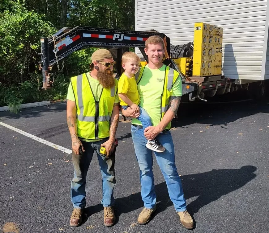 Two men in bright yellow vests standing together, one holding a young child. They are in a parking lot with a truck visible in the background.