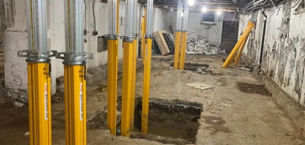 Construction site with metal props supporting the ceiling, and an unfinished concrete floor with a large hole in it.