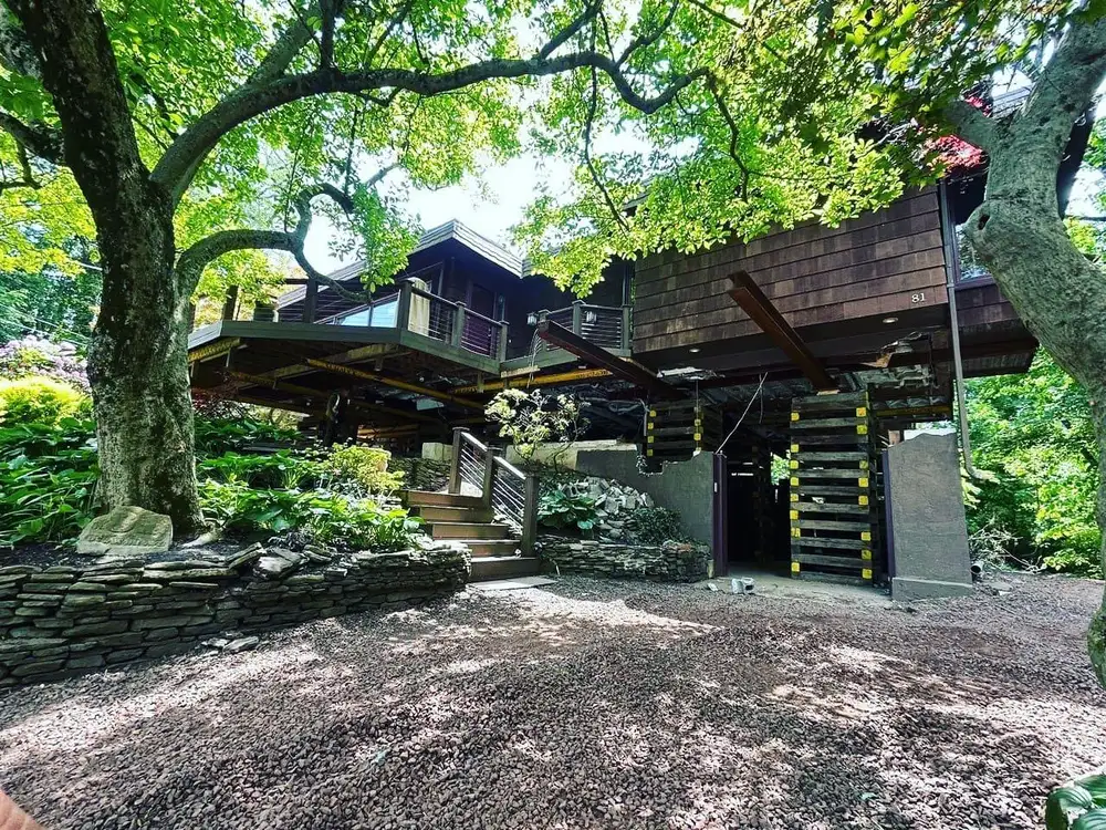 A house surrounded by lush greenery, featuring a unique architectural design with wooden elements and stone landscaping.