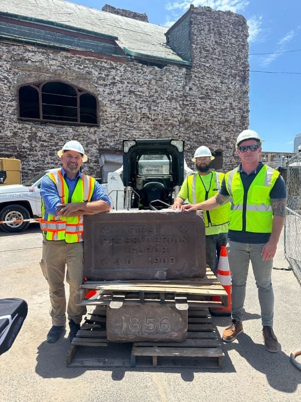 Three construction workers in hard hats and safety vests posing outside next to a large stone block engraved with "FIRST PRESBYTERIAN CHURCH AD 1909" and a lower block marked "1856," with a historic stone building in the background.