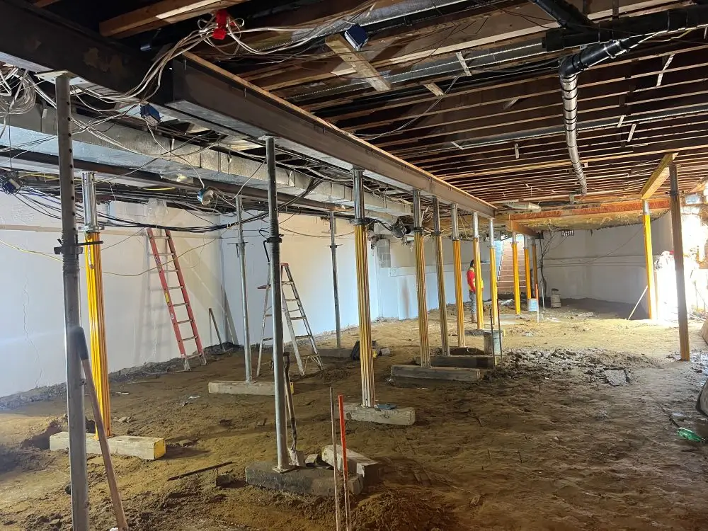 An open basement or crawl space under construction with an unpaved dirt floor, featuring a series of adjustable yellow and silver steel shoring jacks supporting a long steel I-beam to reinforce the overhead wooden floor joists.