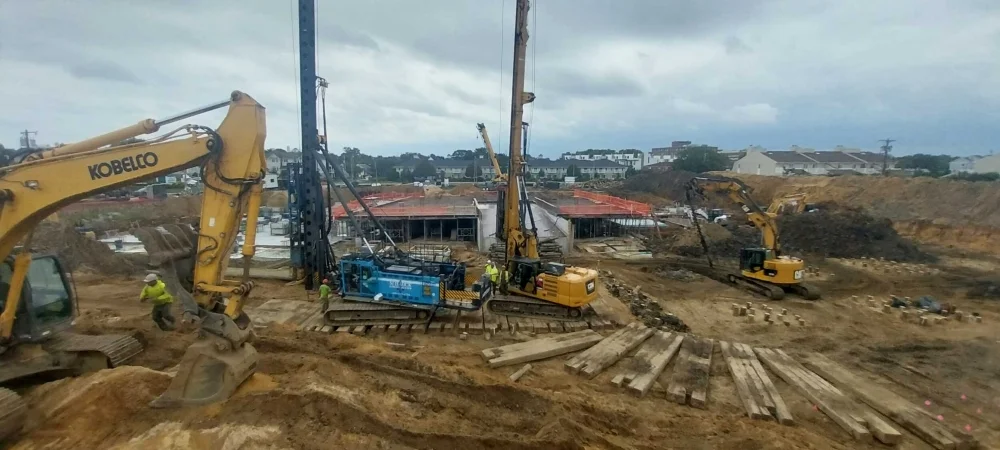 A busy construction site with multiple heavy machines, including yellow excavators and piling drill rigs operating on dirt ground with timber mats, preparing a deep foundation under an overcast sky.