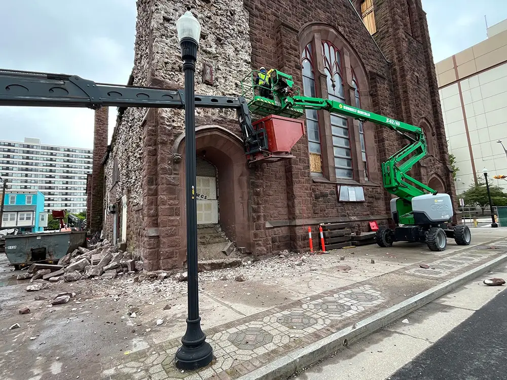 A green Sunbelt Rentals boom lift with a worker in the basket inspects the exterior wall of a historic brown stone church under construction or repair. Debris and broken stones litter the sidewalk near the entrance, with a street lamp in the foreground and a large dumpster nearby.