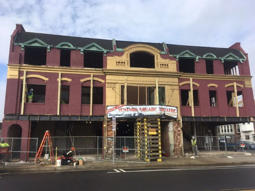 Exterior view of the historic three-story Ventnor Square Theatre building undergoing structural renovation, supported by wooden shoring beams and cribbing stacks behind a chain-link construction fence.