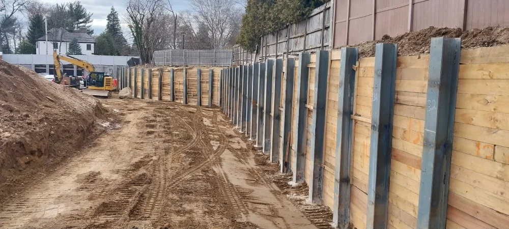 A long soldier pile and lagging retaining wall supporting an excavated dirt embankment at a construction site, with a yellow excavator visible in the background.