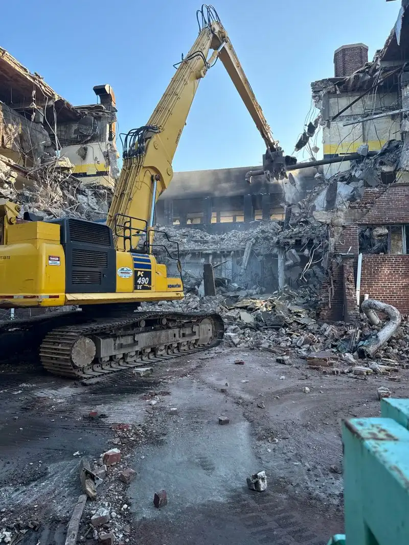 A large yellow Komatsu PC 490 tracked excavator actively tearing down a multi-story brick building on a commercial demolition site filled with concrete rubble and debris.