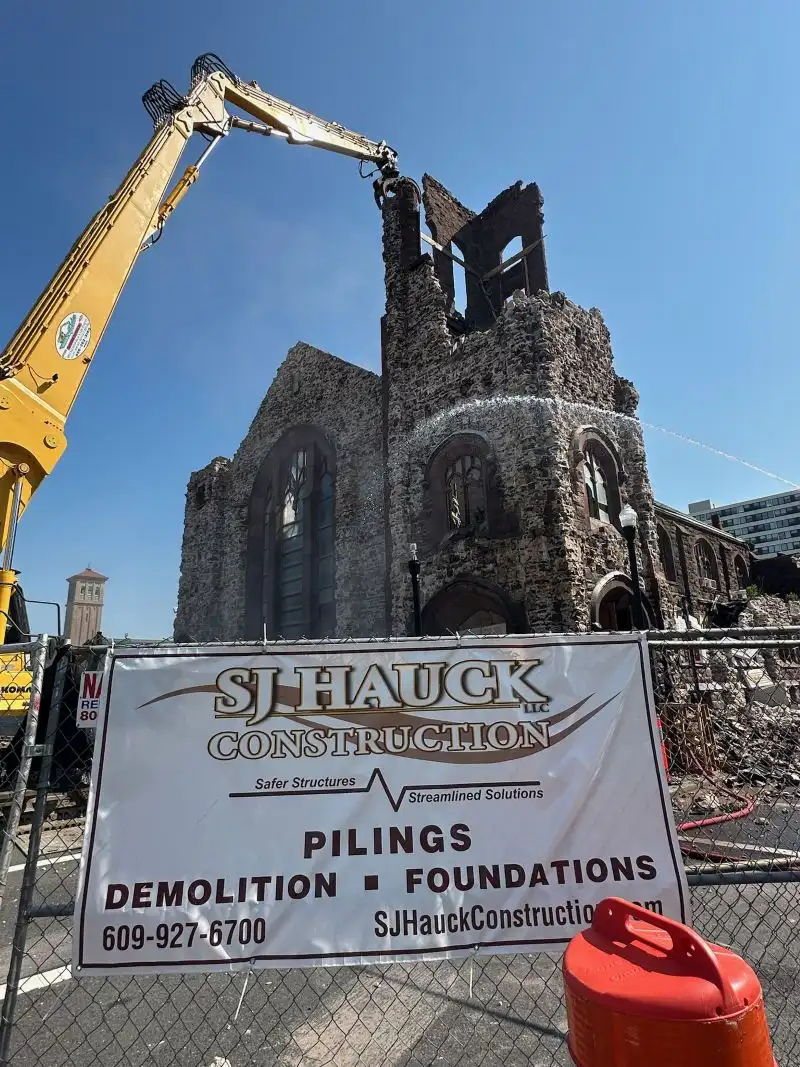 A large yellow excavator actively demolishing a historic stone church building behind a construction fence featuring a prominent "S.J. Hauck Construction" sign advertising pilings, demolition, and foundations.